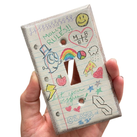 Hand holding a light switch cover with colorful doodles and text.